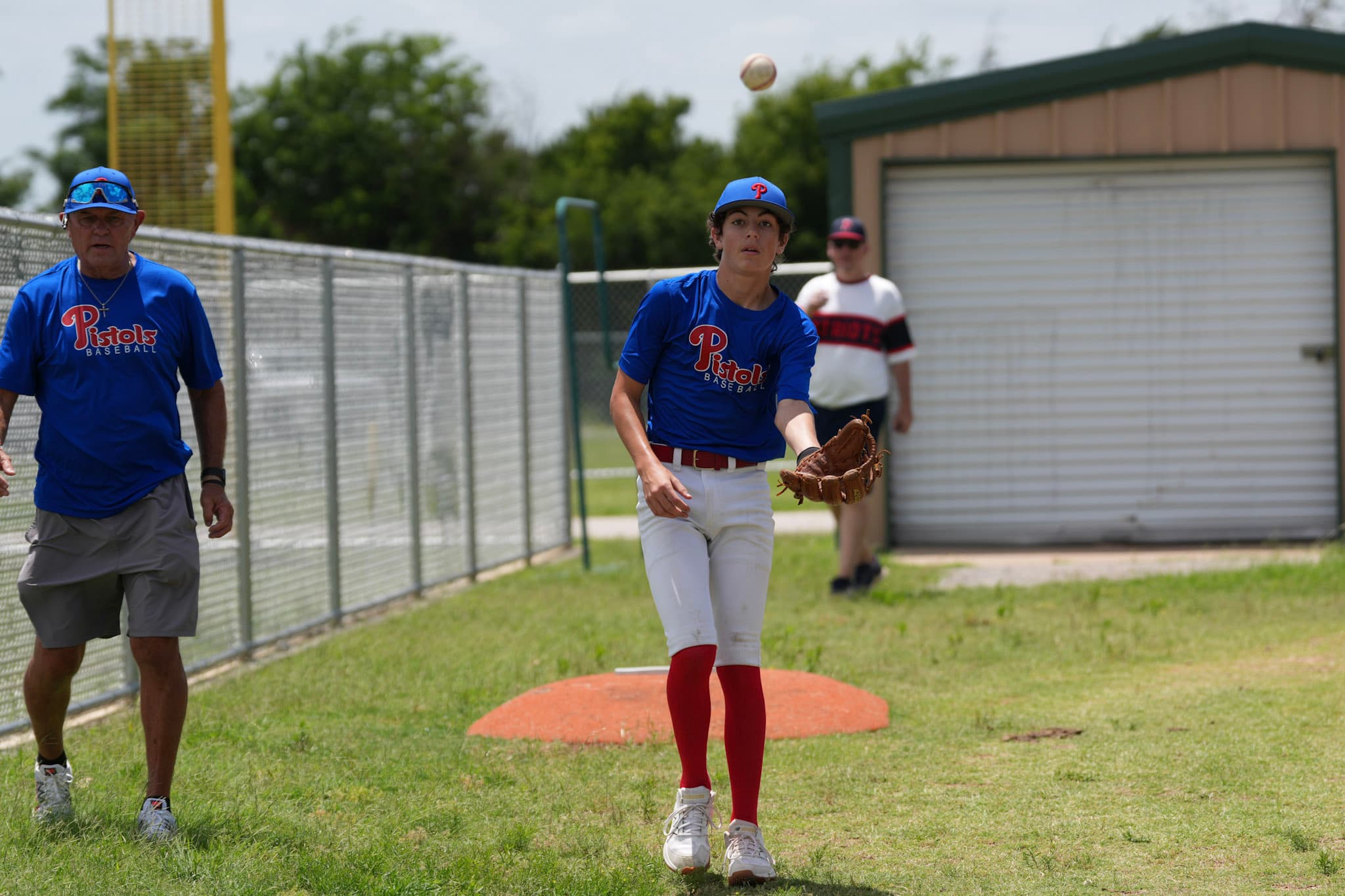 A coach watching a pitcher warm up along the fence line